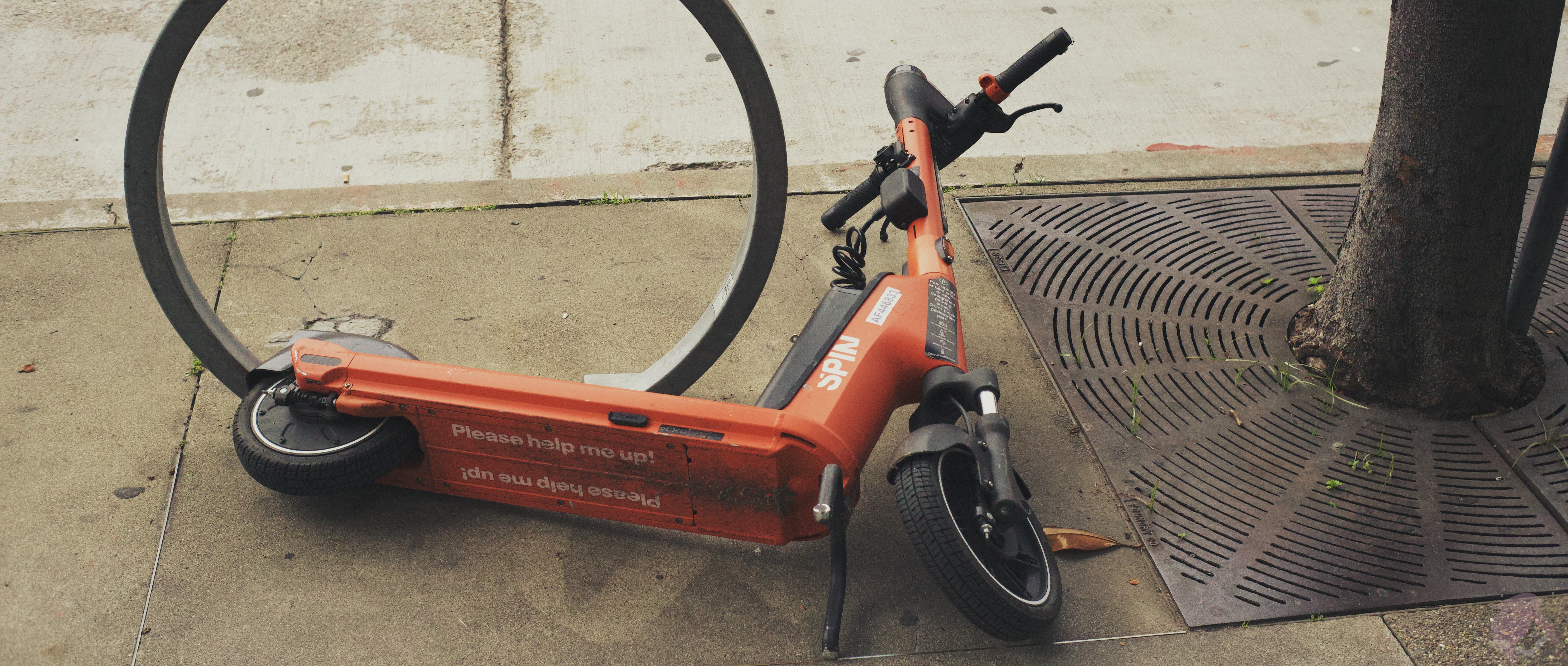 An image of A red electric scooter on its side with the label 'Please help me up' on it.