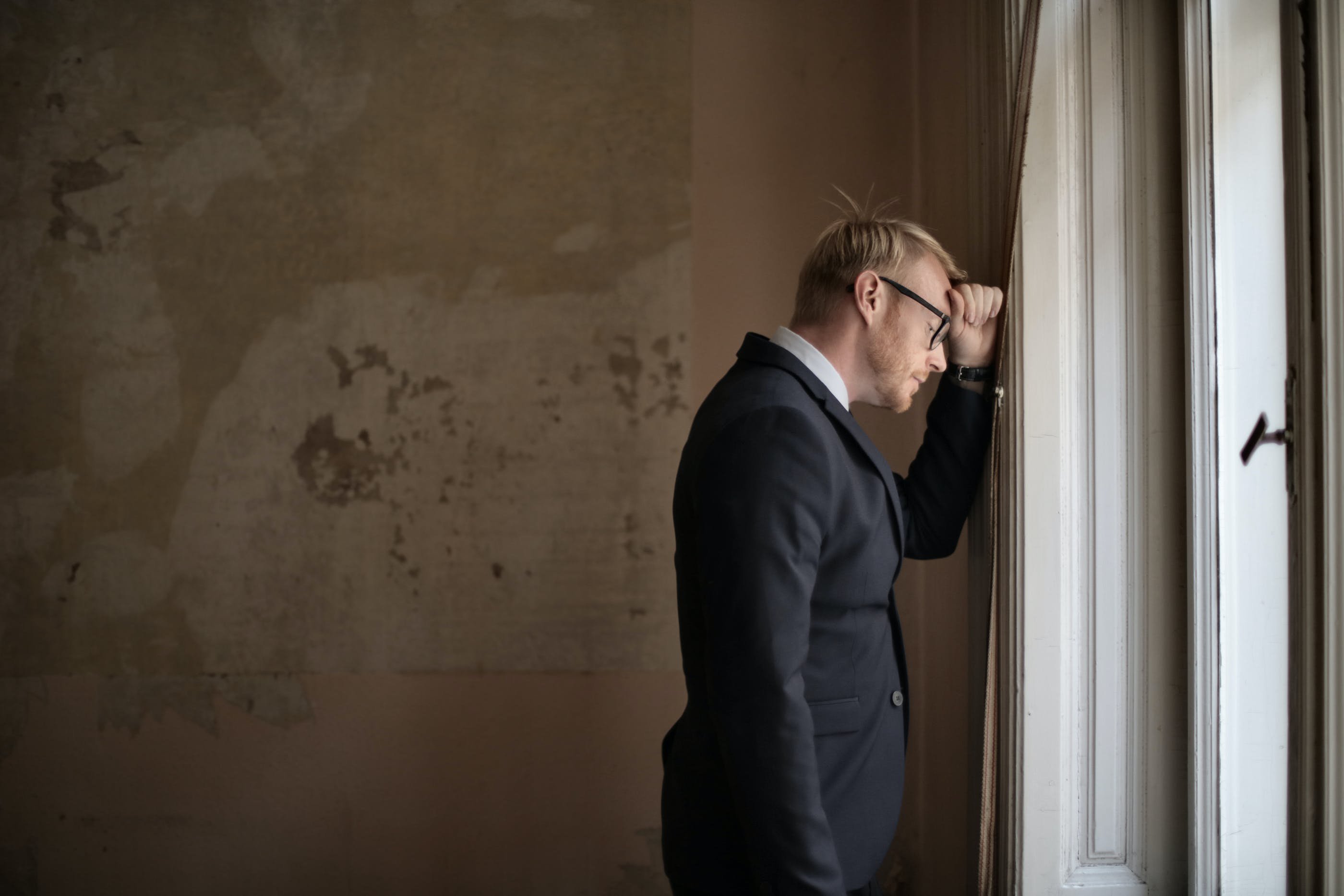 An image of A forlorn business man resting his head on a brown wall next to a window.